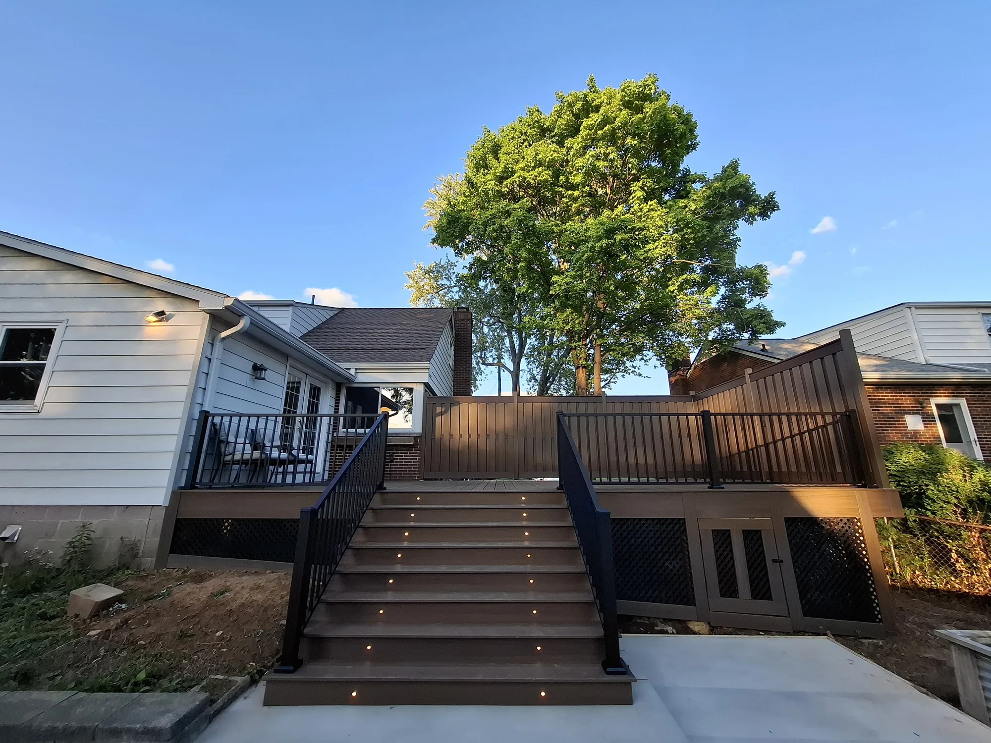 Wide brown composite deck with black railing and lit cascading steps down to a concrete pad at a white sided home