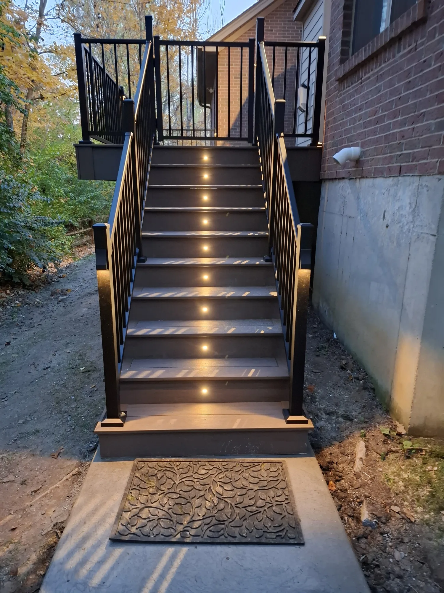 Dark composite staircase with lights set into each riser leading up to a deck against a brick home