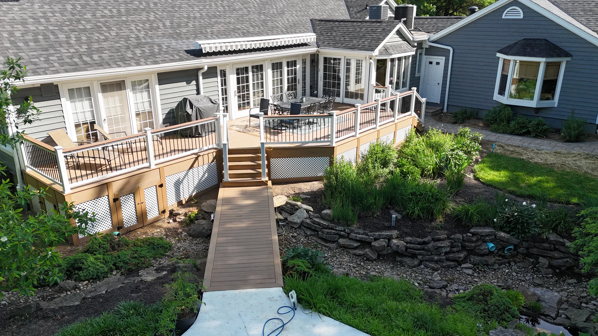 Aerial view of a multi level composite deck with white railing and a walkway crossing a landscaped rock garden at a blue home
