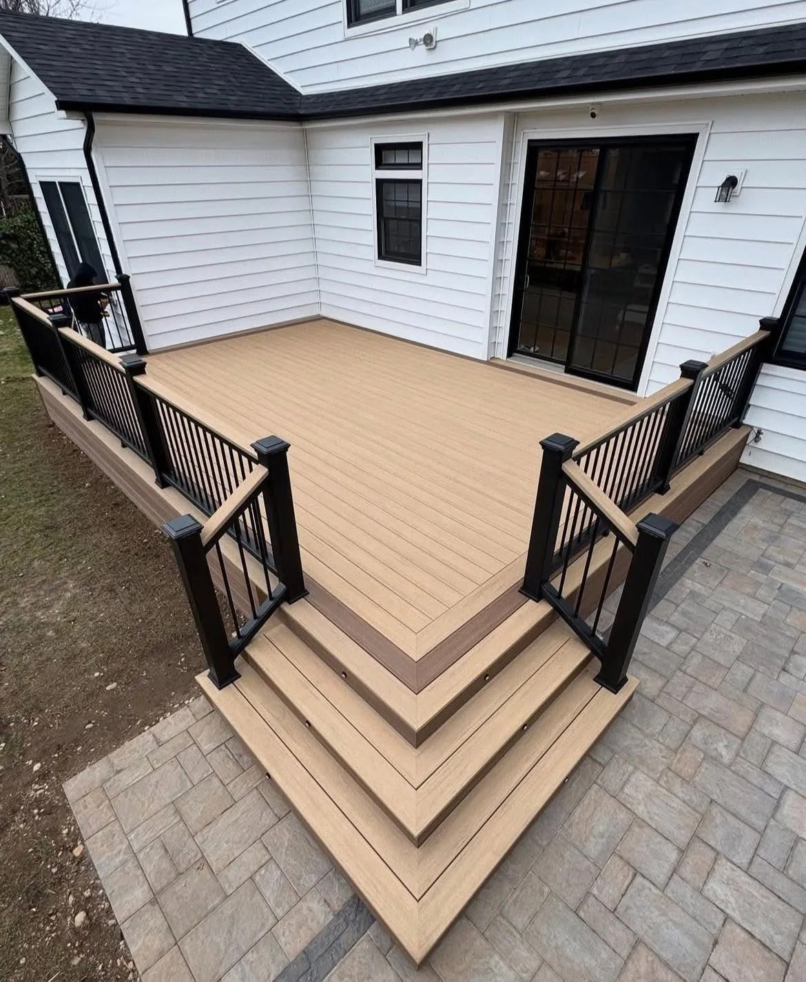Tan composite deck with black railing and wide cascading corner steps down to a paver patio at a white home