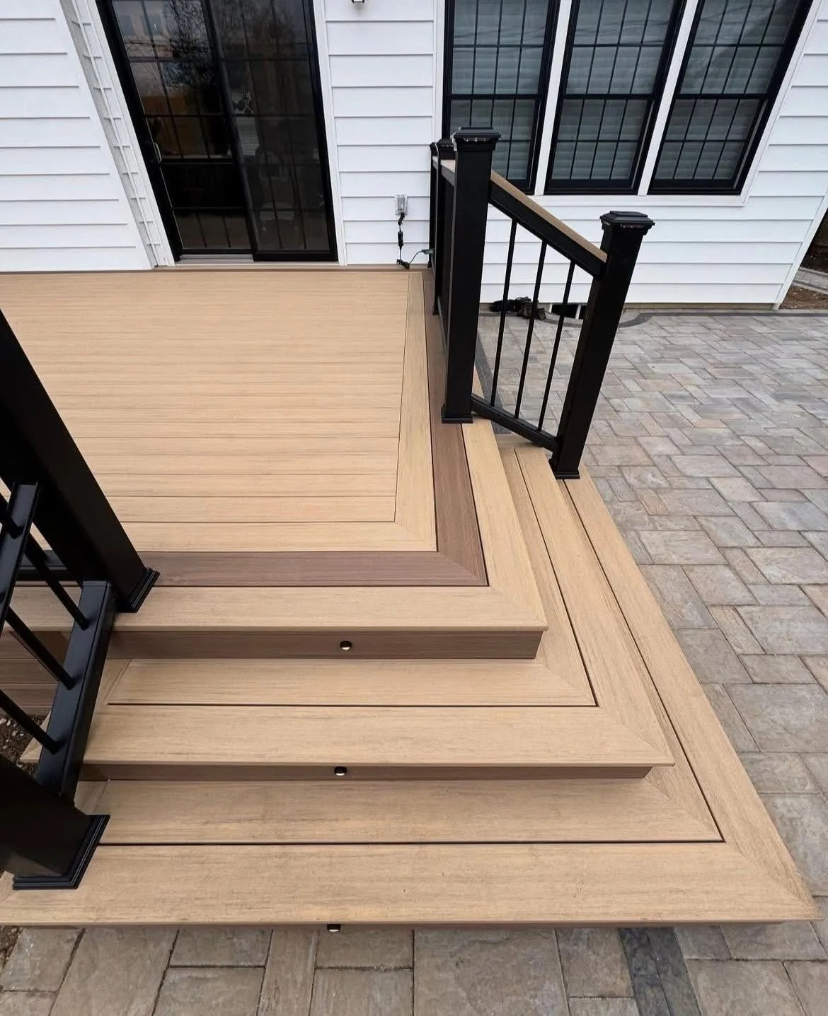 Tan composite cascading steps with a black picture frame border and black railing over a paver patio