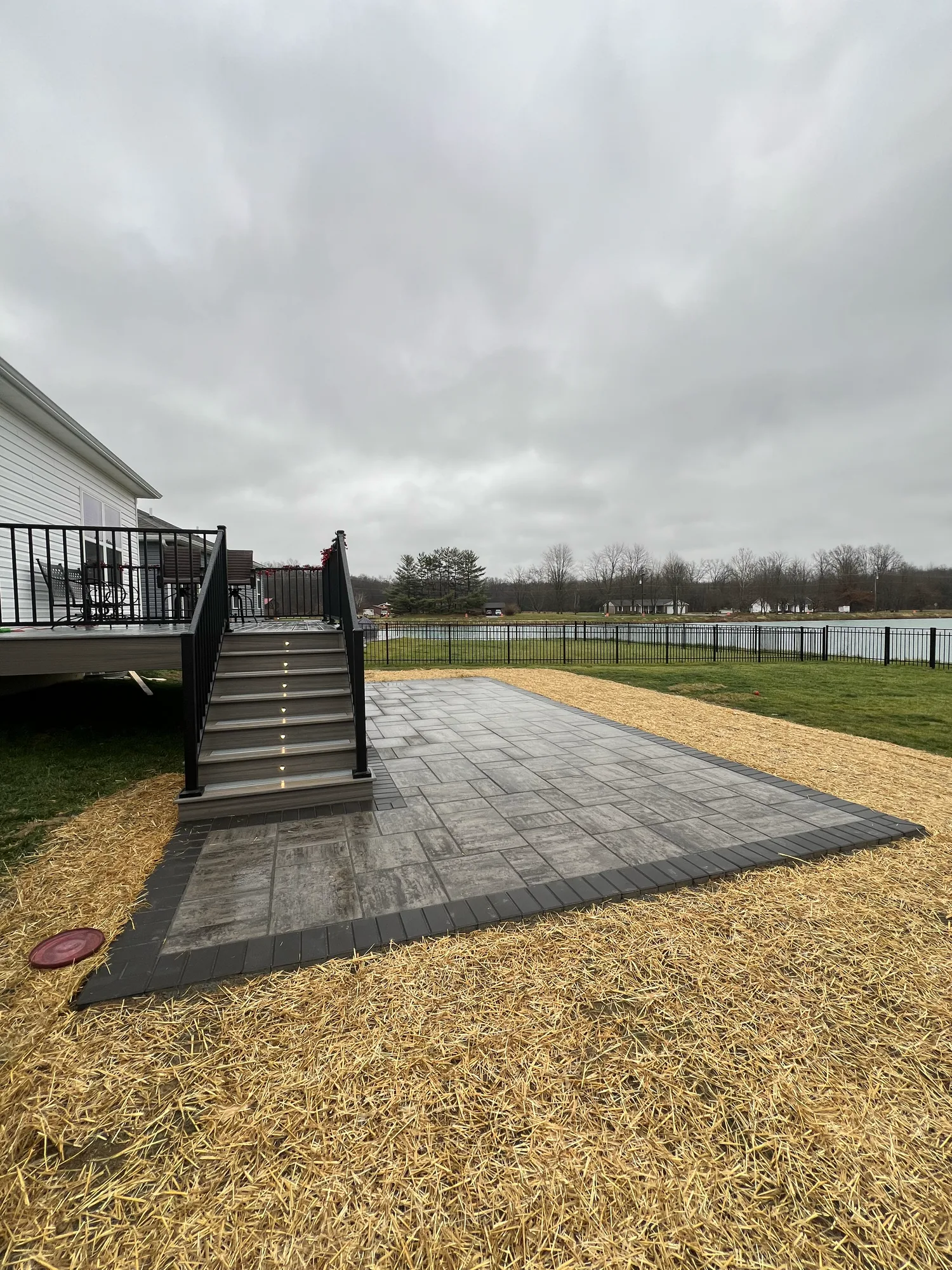 Grey composite deck with black railing and steps beside a strawed yard looking out to water