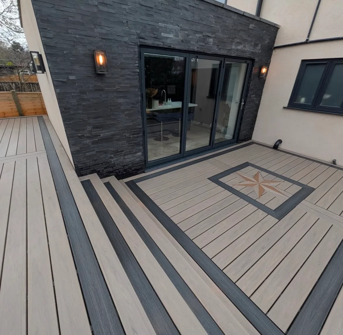 Composite deck with a compass star inlay and dark border boards outside a modern dark stone home