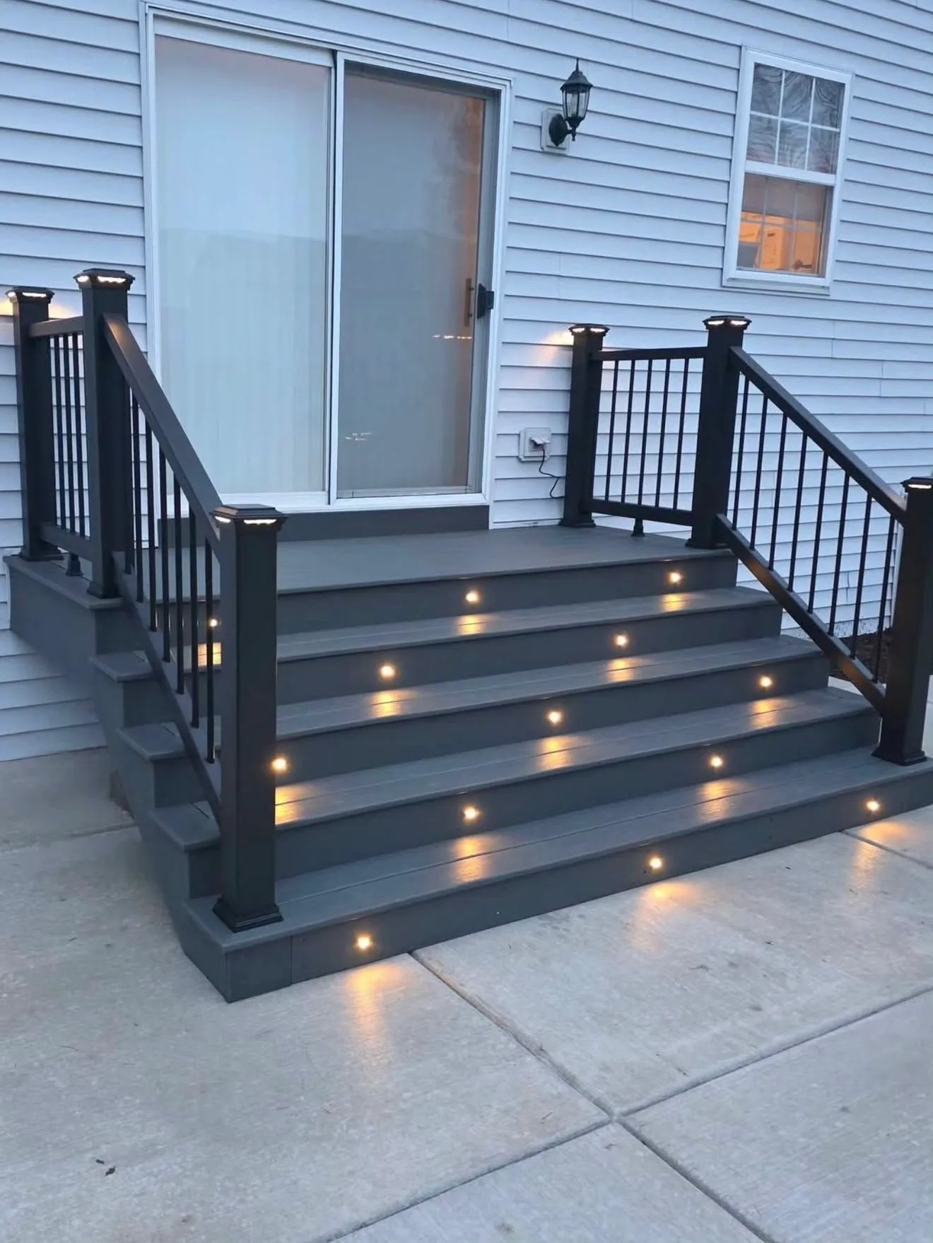 Dark composite deck steps with lights set into each riser and black railing at a blue sided home at dusk