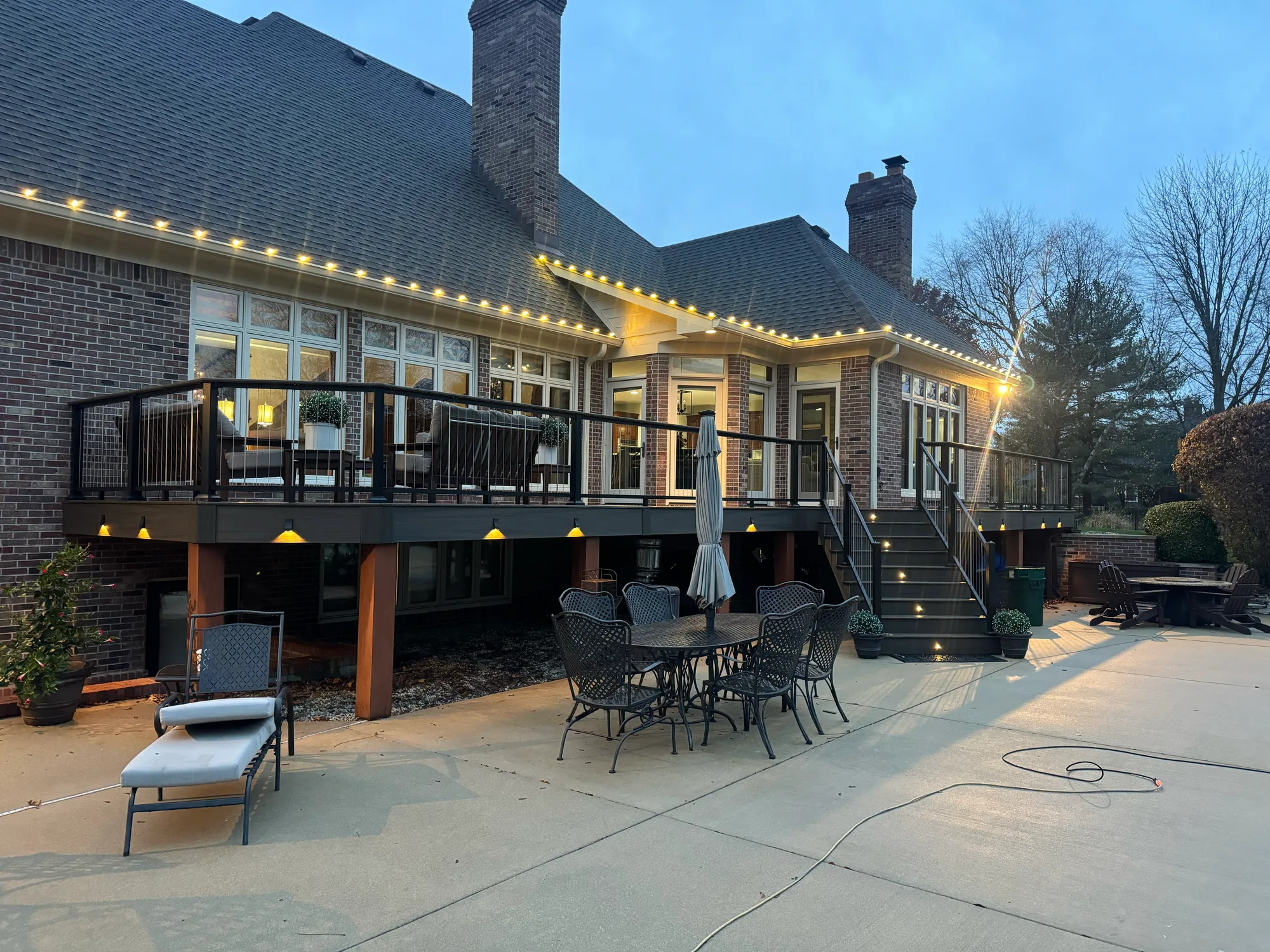 Elevated composite deck lit with string lights at dusk above a concrete patio with dining furniture at a large brick home