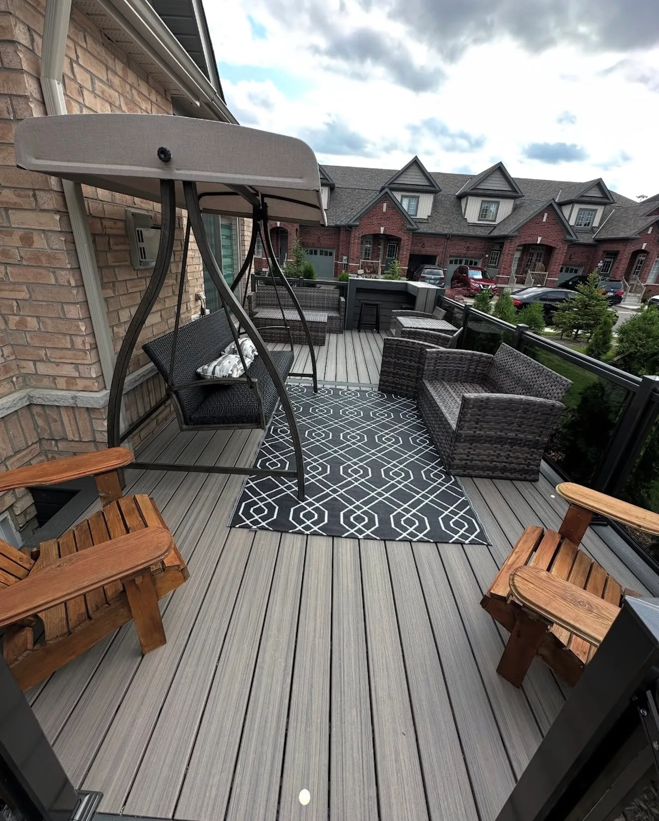 Grey composite deck with a swing, wooden chairs and a patterned rug behind a row of townhomes