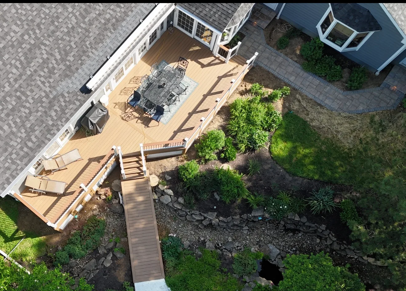 Aerial view of a paver patio and composite deck with a dining set and a walkway crossing a rock garden