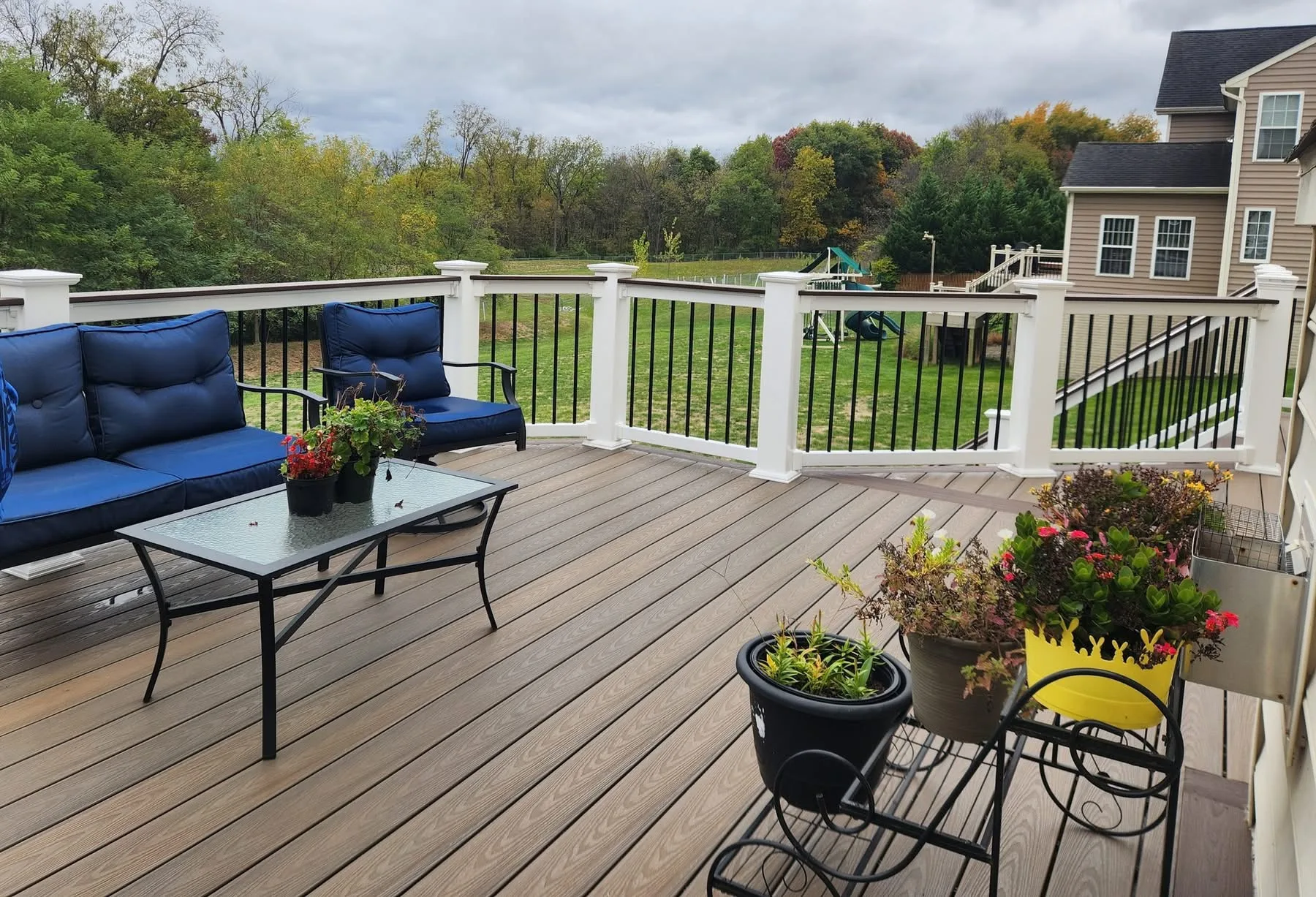 Brown composite deck with blue cushioned furniture, potted plants and white railing overlooking a park
