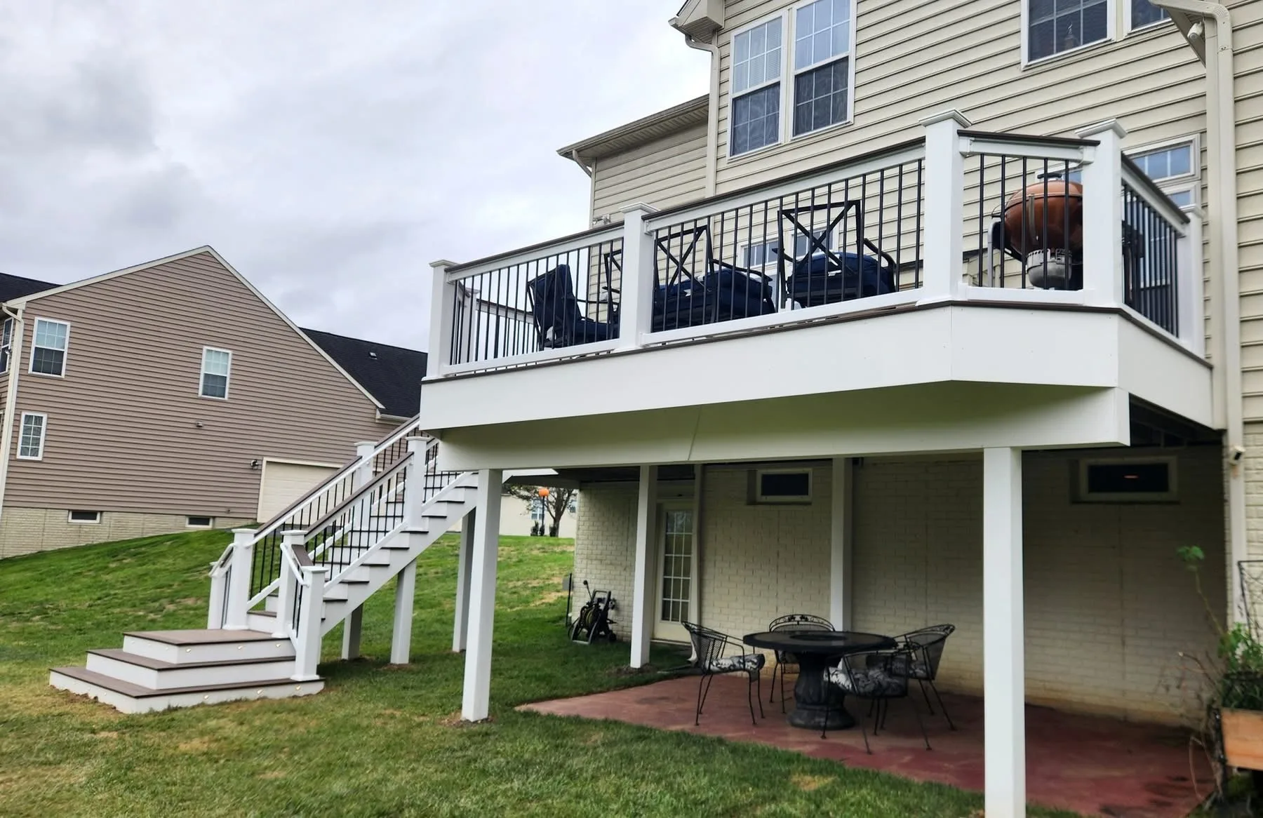 Elevated composite deck with black railing and a white staircase above a fire pit patio at a tan home