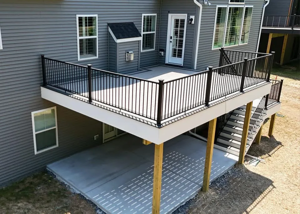 Raised grey composite deck with black railing on posts above a concrete patio at a grey home